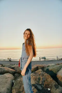 Улыбающаяся девушка с рыжими волосами стоит на камнях у моря на закате