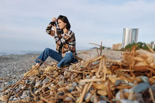 Девушка в клетчатой рубашке фотографирует на каменистом берегу
