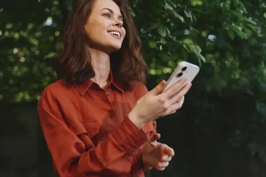 Улыбающаяся девушка в красной рубашке смотрит в смартфон на фоне зелени