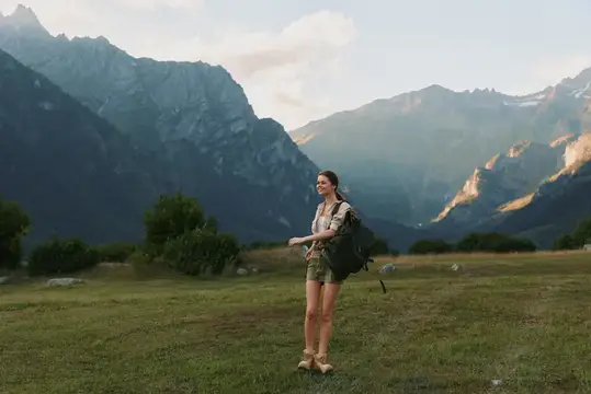 Улыбающаяся девушка с рюкзаком стоит на горном лугу