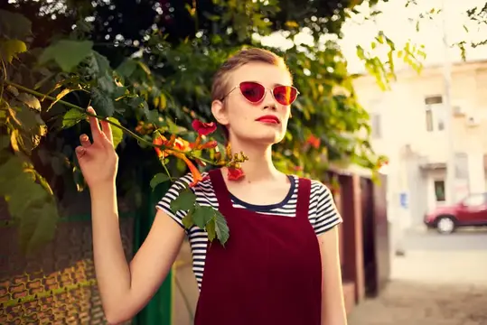 Молодая женщина в красных очках и бордовом сарафане у куста