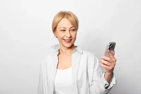 Пожилая женщина улыбается со смартфоном, портрет на сером фоне