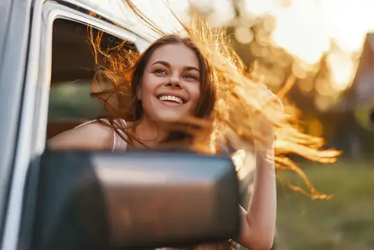 Радостная молодая женщина с развевающимися волосами улыбается из окна автомобиля во время путешествия, окруженная яркой атмосферой заката