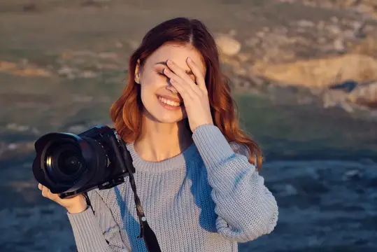 Рыжеволосая девушка с фотокамерой смеётся, закрыв лицо рукой на закате