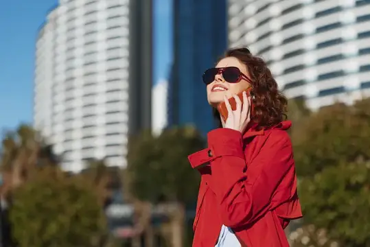 Улыбающаяся кудрявая женщина в красном плаще говорит по телефону на фоне высоток