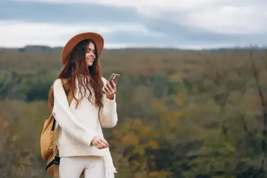 Рыжеволосая девушка в шляпе смотрит в телефон на фоне осеннего поля
