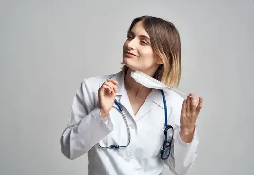 Молодая женщина-врач в белом халате держит медицинскую маску и улыбается