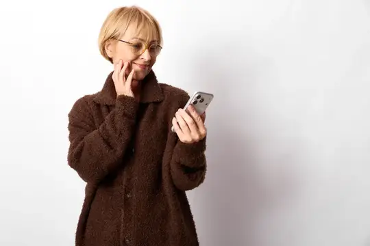Блондинка в очках и коричневом пальто смотрит в смартфон на белом фоне