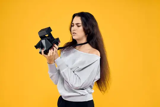 Девушка с длинными волосами держит чёрную фотокамеру на жёлтом фоне