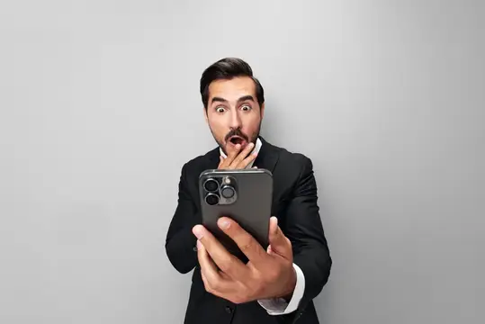Удивлённый мужчина в костюме смотрит в смартфон на сером фоне