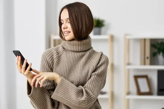Улыбающаяся девушка с каре в свитере смотрит в смартфон дома