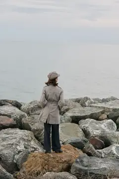 Женщина в плаще и кепке стоит на камнях у моря