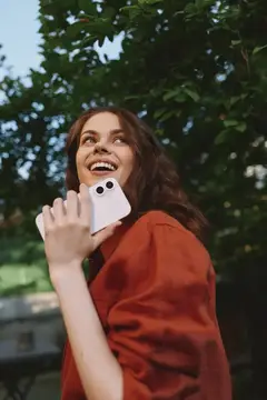 Счастливая женщина на улице разговаривает по смартфону в окружении деревьев и зелени, что передаёт расслабленный образ жизни, искренний момент и повседневную связь