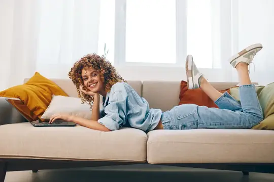 Кудрявая девушка лежит на диване с ноутбуком и улыбается