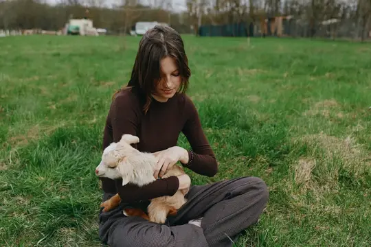 Естественная обстановка показывает женщину с козлёнком в расслабленный момент, расслабленная поза, лёгкий ветерок, зелёный фон, деревенская атмосфера, мирная связь между человеком и животным…