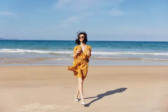 Улыбающаяся темноволосая женщина в жёлтом платье бежит по пляжу