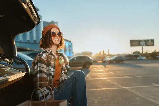 Улыбающаяся девушка в солнечных очках сидит у открытого багажника автомобиля