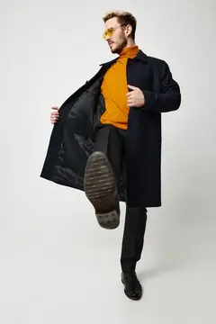 Модный мужчина танцует на светлом фоне, одетый в чёрное пальто, оранжевый свитер и брюки