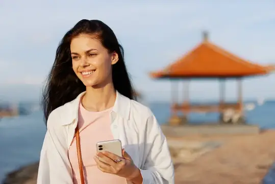 Женщина, улыбающаяся блогер-фрилансер с телефоном в руке, отдыхает, гуляя по пляжу у океана на закате на Бали, счастливое путешествие и отпуск, мобильная связь, интернет онлайн