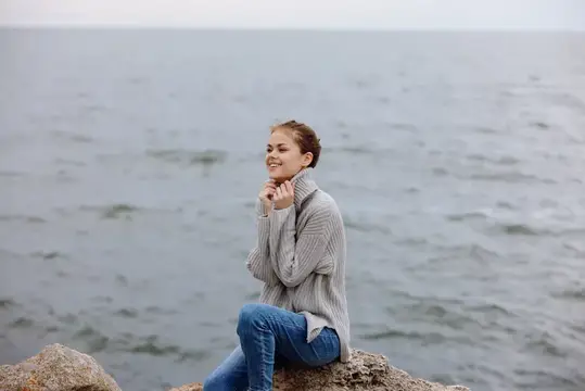 Молодая женщина в сером свитере сидит на камнях у моря