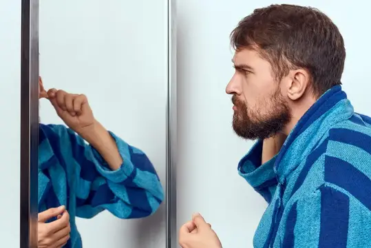Бородатый мужчина в синем халате смотрит на своё отражение в зеркале