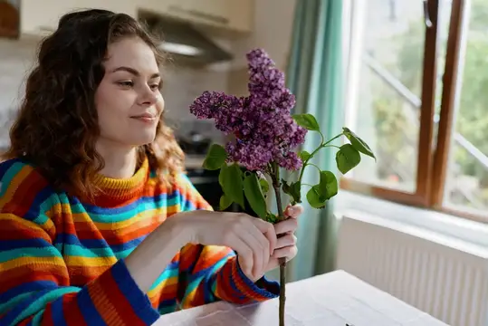 Молодая женщина с сиренью в руках у окна на кухне