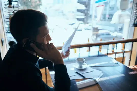 Мужчина говорит по телефону с бумагами у окна кафе