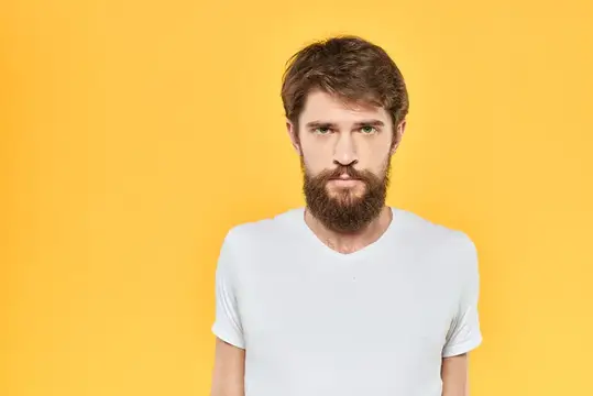 Бородатый мужчина в белой футболке демонстрирует эмоции, жестикулирует руками и выражает мимикой на жёлтом фоне