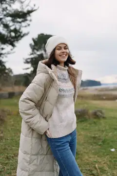 Зимняя мода, на открытом воздухе, уютный наряд, тёплое пальто, повседневный стиль, улыбающаяся женщина в вязаном свитере, шапке-бини и джинсах стоит на открытом ландшафте под облачным небом…