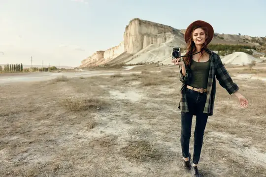Женщина-фотограф в клетчатой рубашке и шляпе снимает природу на камеру, находясь на открытом поле