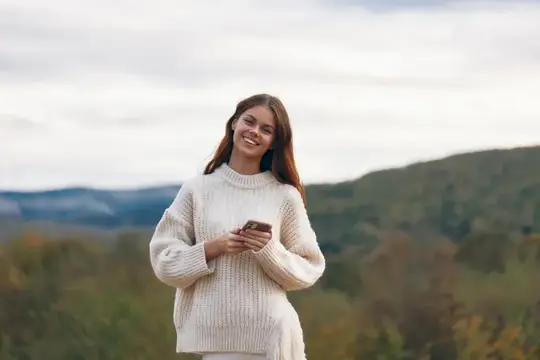 Улыбающаяся девушка в белом свитере с телефоном на фоне гор
