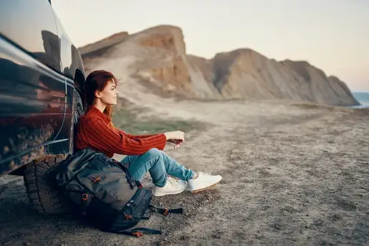 Рыжеволосая девушка отдыхает у машины на фоне гор