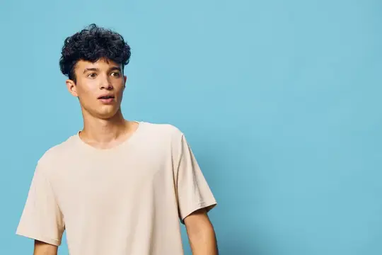 Кудрявый парень в бежевой футболке удивлённо смотрит в сторону на голубом фоне
