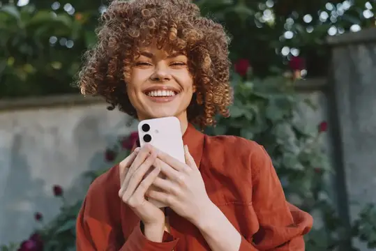 Кудрявая девушка смеётся, глядя в смартфон на улице