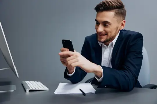 Улыбающийся мужчина в костюме смотрит в смартфон за рабочим столом