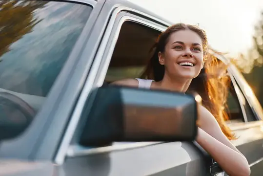 Счастливая молодая женщина с развевающимися волосами высунулась из окна автомобиля, широко улыбаясь на фоне живописного заката, что передает суть приключений и свободы в путешествии