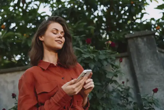 Молодая женщина стоит среди зелени в ржаво-коричневой рубашке, смотрит на смартфон с расслабленным выражением лица