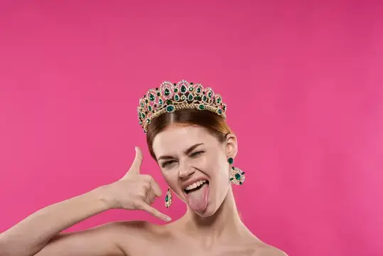 Молодая женщина в короне с высунутым языком на розовом фоне