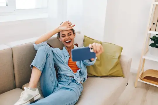 Радостная женщина отдыхает на уютном диване, держит смартфон и улыбается