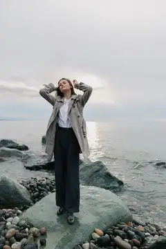 Молодая женщина в плаще стоит на камне у моря с закрытыми глазами