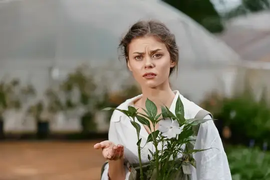 Молодая женщина с недовольным лицом держит саженец в теплице