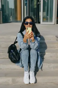 Улыбающаяся темноволосая девушка в джинсовом костюме делает селфи на ступеньках