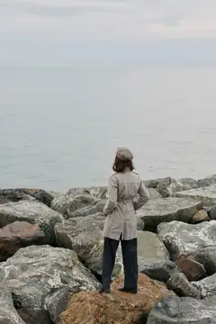 Женщина в пальто и шляпе стоит на камнях у моря, глядя на горизонт в облачный день