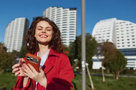 Улыбающаяся кудрявая женщина в красном пальто с телефоном на фоне высоток