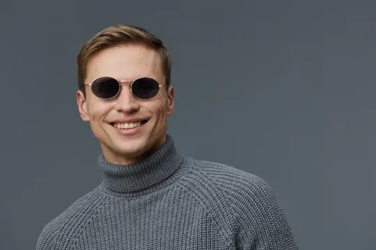 Улыбающийся мужчина с короткими волосами и модными черными солнцезащитными очками, одетый в серую водолазку, уверенно позирует в студии на нейтральном сером фоне