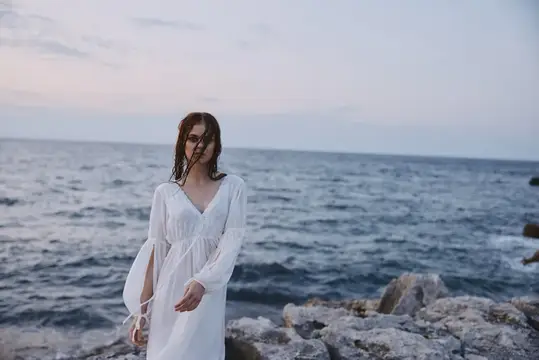 Девушка в белом платье стоит на скалистом берегу моря