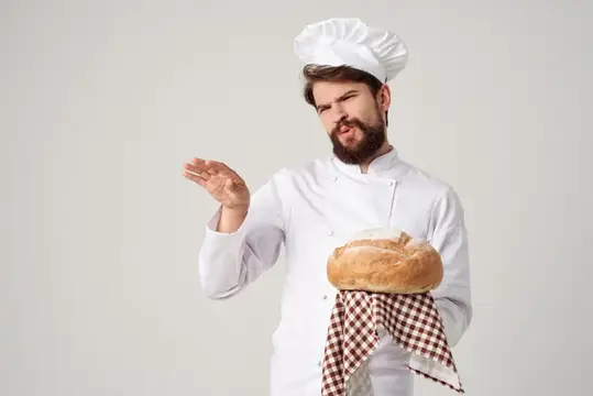 Бородатый пекарь в колпаке показывает рукой на каравай