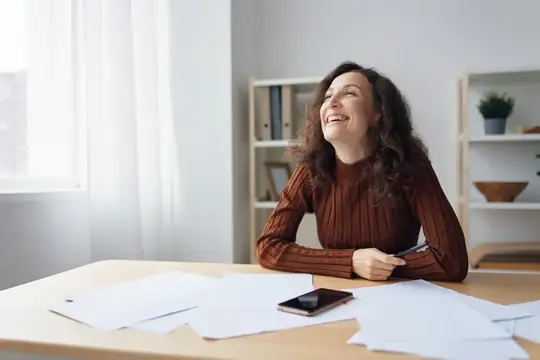 Смеющаяся, счастливая, жизнерадостная, кудрявая, милая леди сидит за столом с бумагами и документами, работает из дома, думает о будущих отпусках, мечтает о выходных и смотрит в сторону на окно