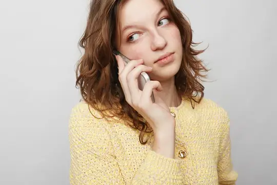 Женщина с вьющимися волосами в желтом вязаном свитере прижимает смартфон к уху, глядя в сторону с задумчивым настроением
