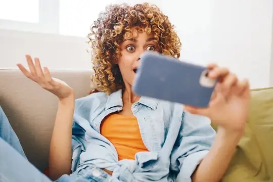 Кудрявая девушка в джинсовой рубашке удивлённо смотрит в смартфон